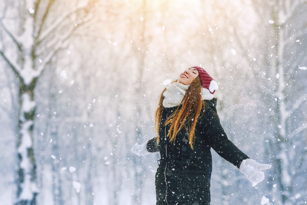 Talvivaatteisiin pukeutunut nainen seisoo ulkona lumisateessa ja näyttää tyytyväiseltä.